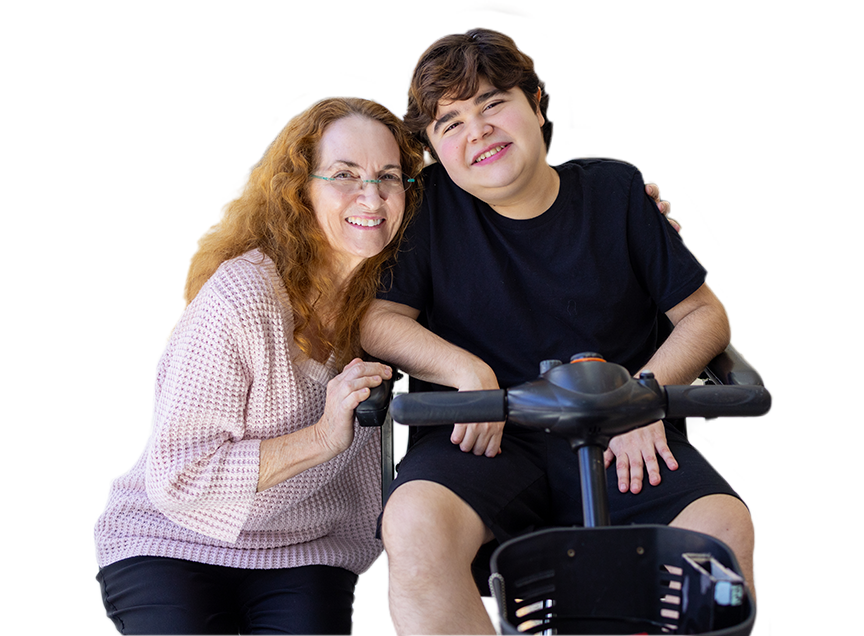 A little boy is wearing all black and is sitting on a bike wheelchair, smiling at the camera and next to him is an older lady wearing a pink shirt with her arms around him.