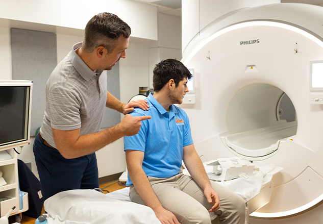 An older man in a grey shirt is pointing towards an MRI machine with his other hand on the shoulder of a younger man, wearing a blue shirt, who is sitting on the bed, also looking towards the MRI machine.